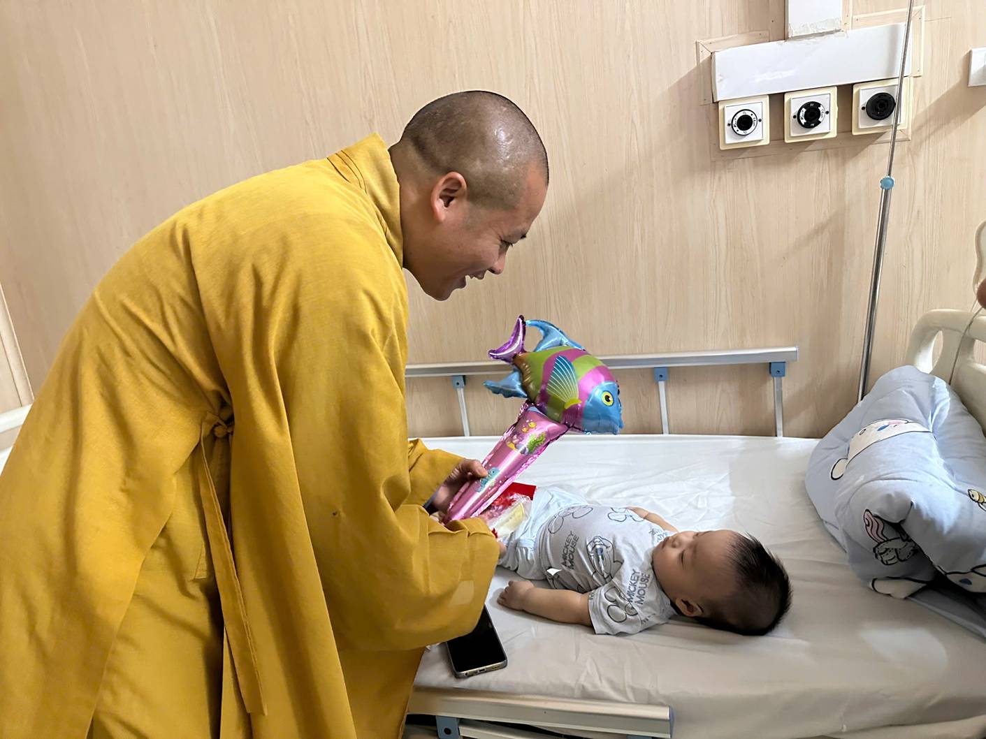 A person in a yellow robe holding a balloon next to a baby lying on a bed

AI-generated content may be incorrect.